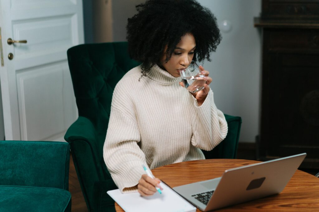 pexels photo 6986459 6986459 A woman with afro hair working from home, taking notes and drinking water.